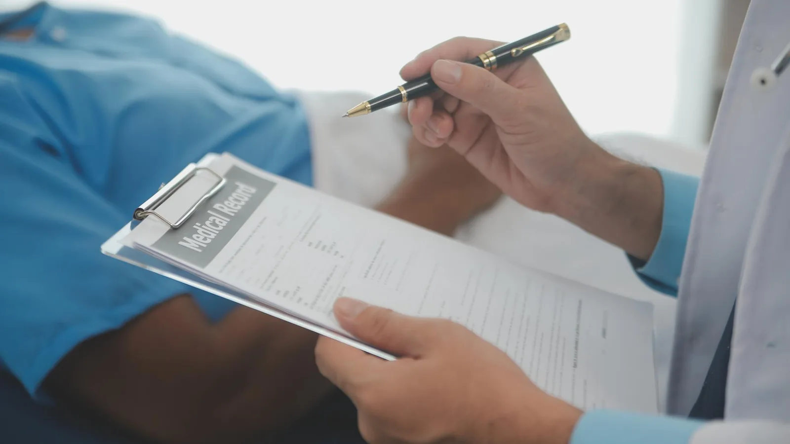 A healthcare professional holding a clipboard with a medical record form while preparing to write with a pen, with a patient in blue scrubs visible in the background.