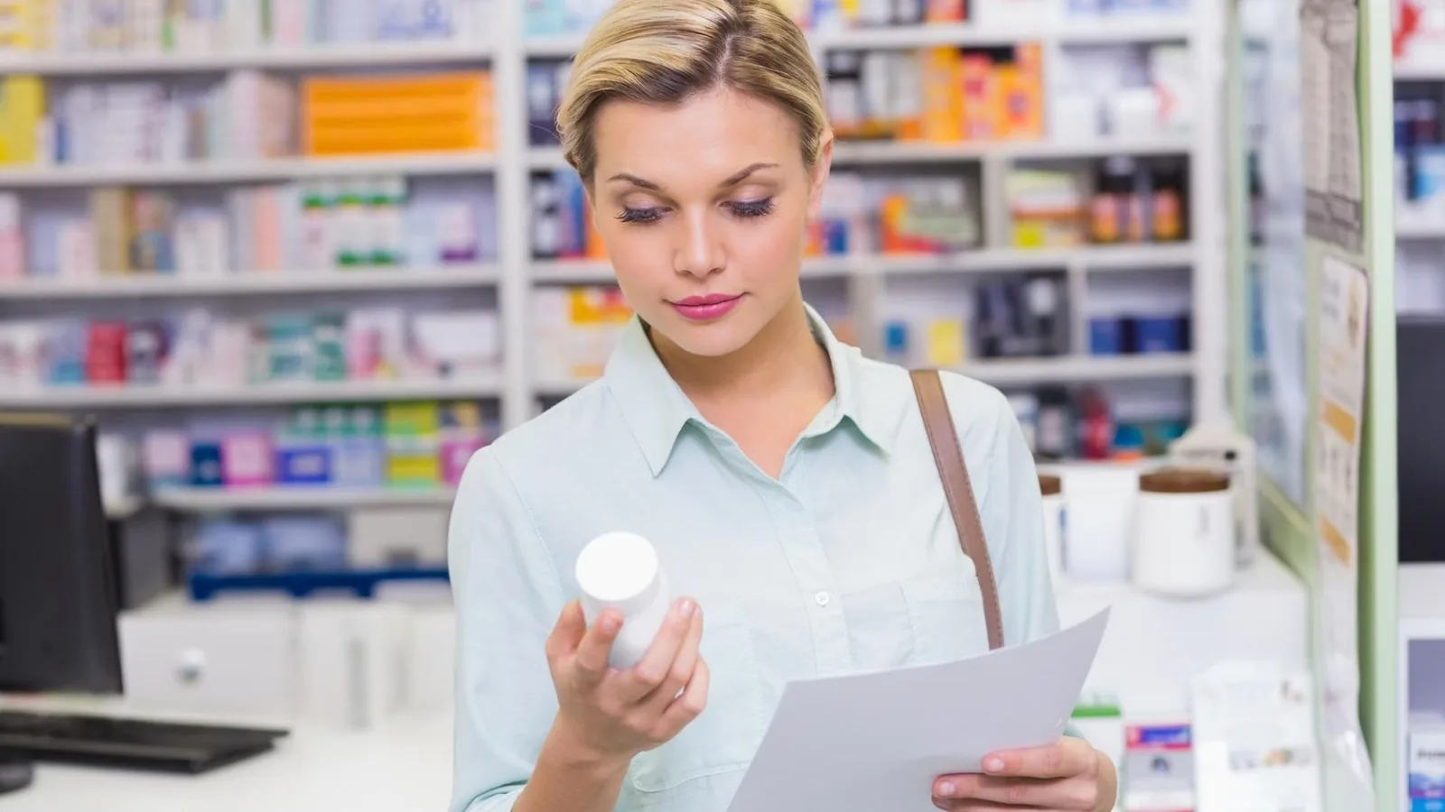 A blonde woman in a pharmacy holding a white medicine bottle and a piece of paper, looking down at them.